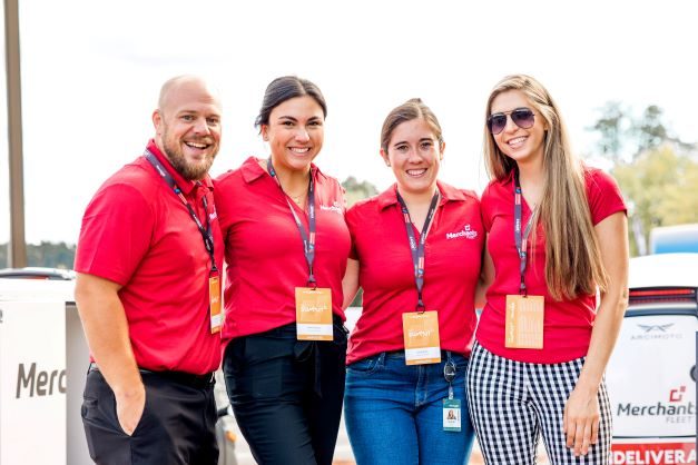 Four people in red shirts smiling, wearing name tags and standing outdoors in front of a Merchant logo.