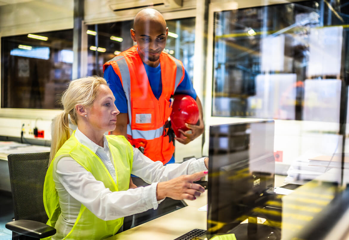 Two workers in safety vests are at a computer. One is seated and pointing at the screen, the other is standing and observing.