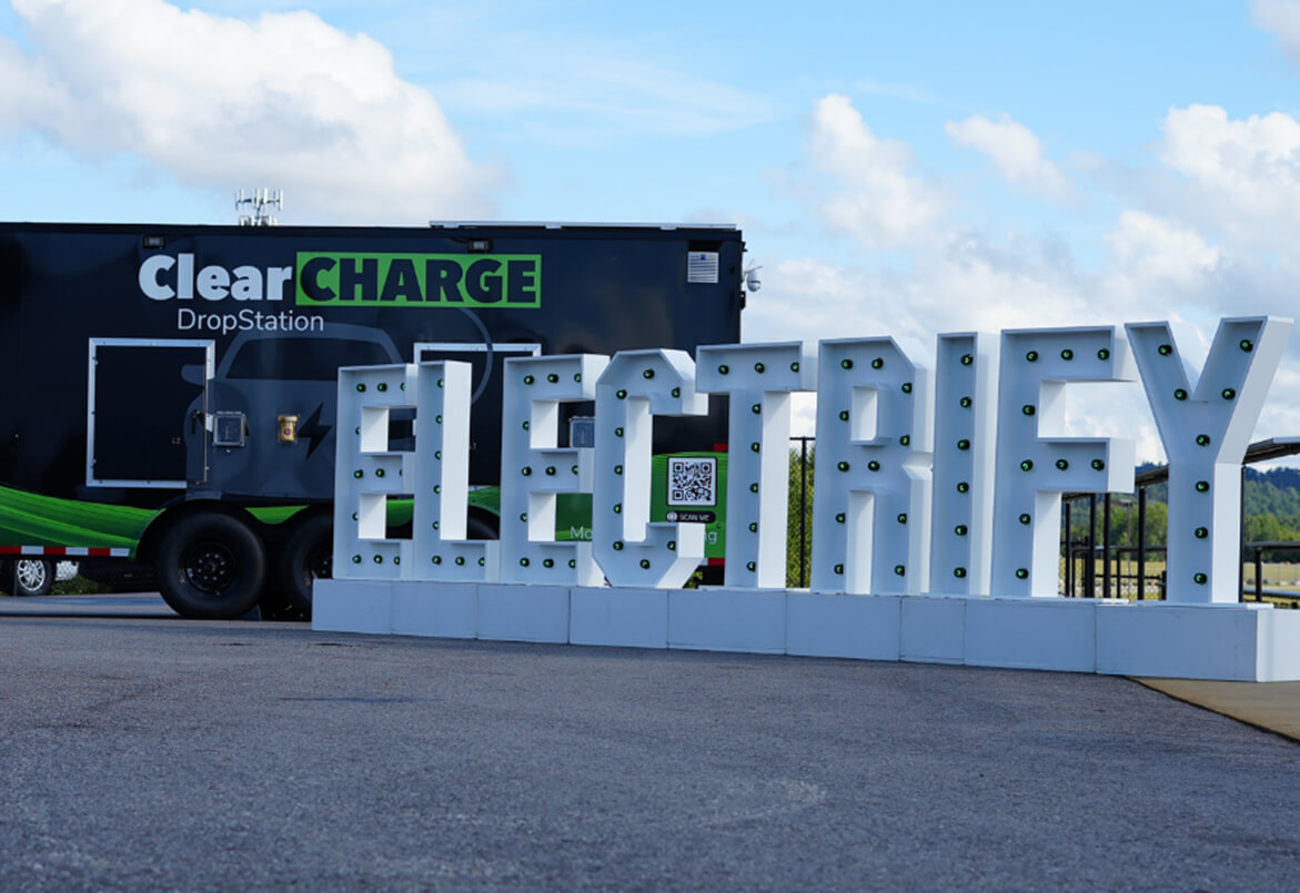 Large white 3D letters spell "ELECTRIFY" beside a black trailer with "ClearCHARGE DropStation" on it.