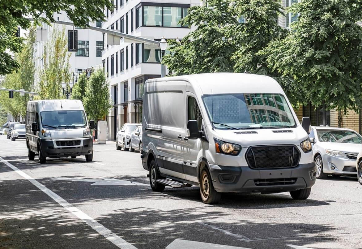 White cargo vans driving on a city street lined with trees and buildings on a sunny day.
