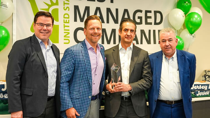 Four men in suits standing in front of a banner, one holding a glass award.