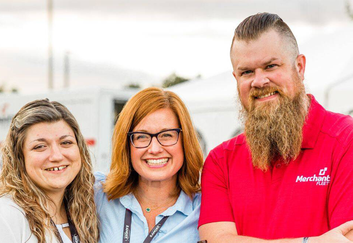 Three smiling people outdoors; one wearing a red shirt, the others in light-colored tops.