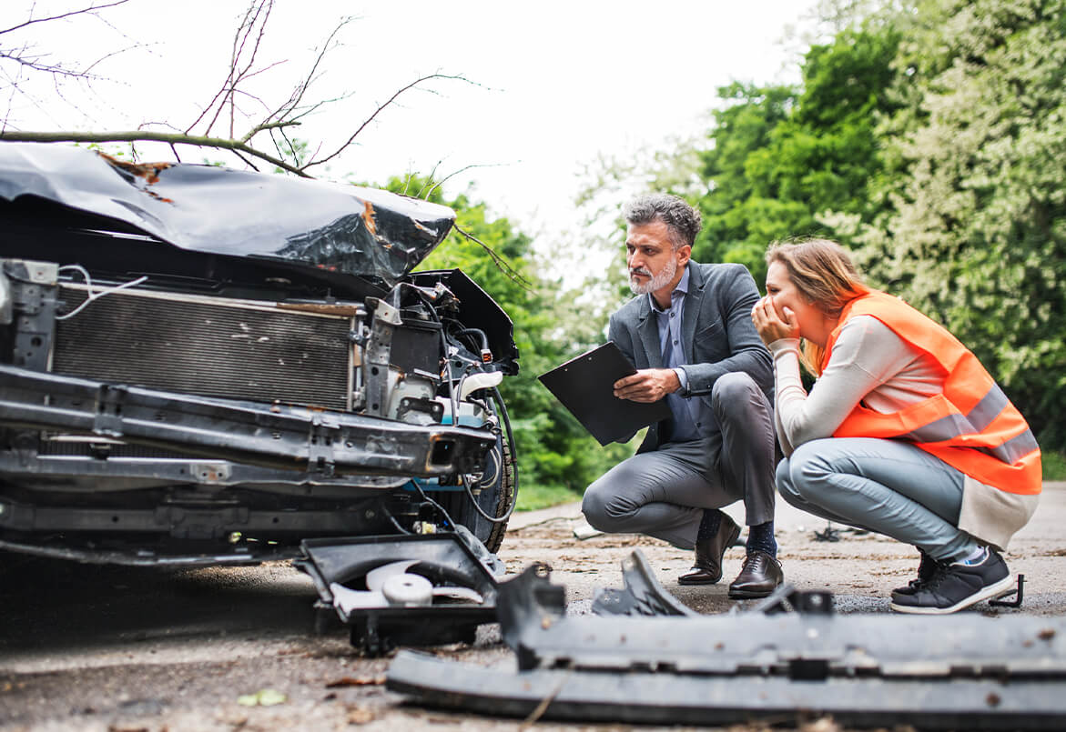 A person in a suit and a person in an orange vest examine a damaged car on a road.