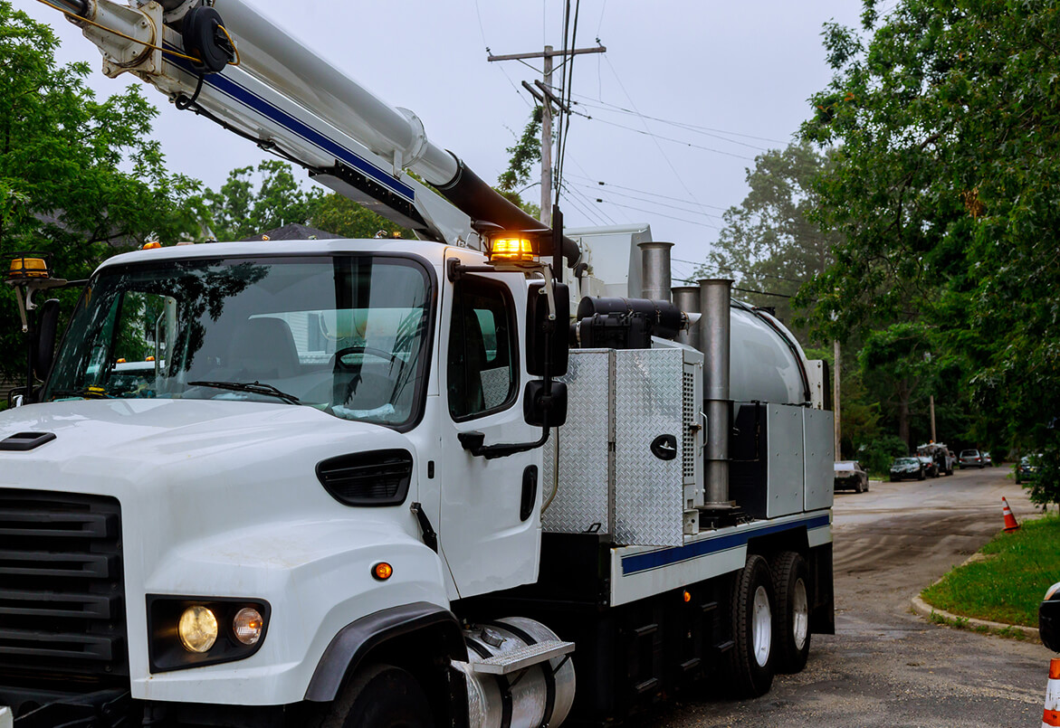 White utility truck with crane arm parked on a suburban street with trees and power lines in the background.