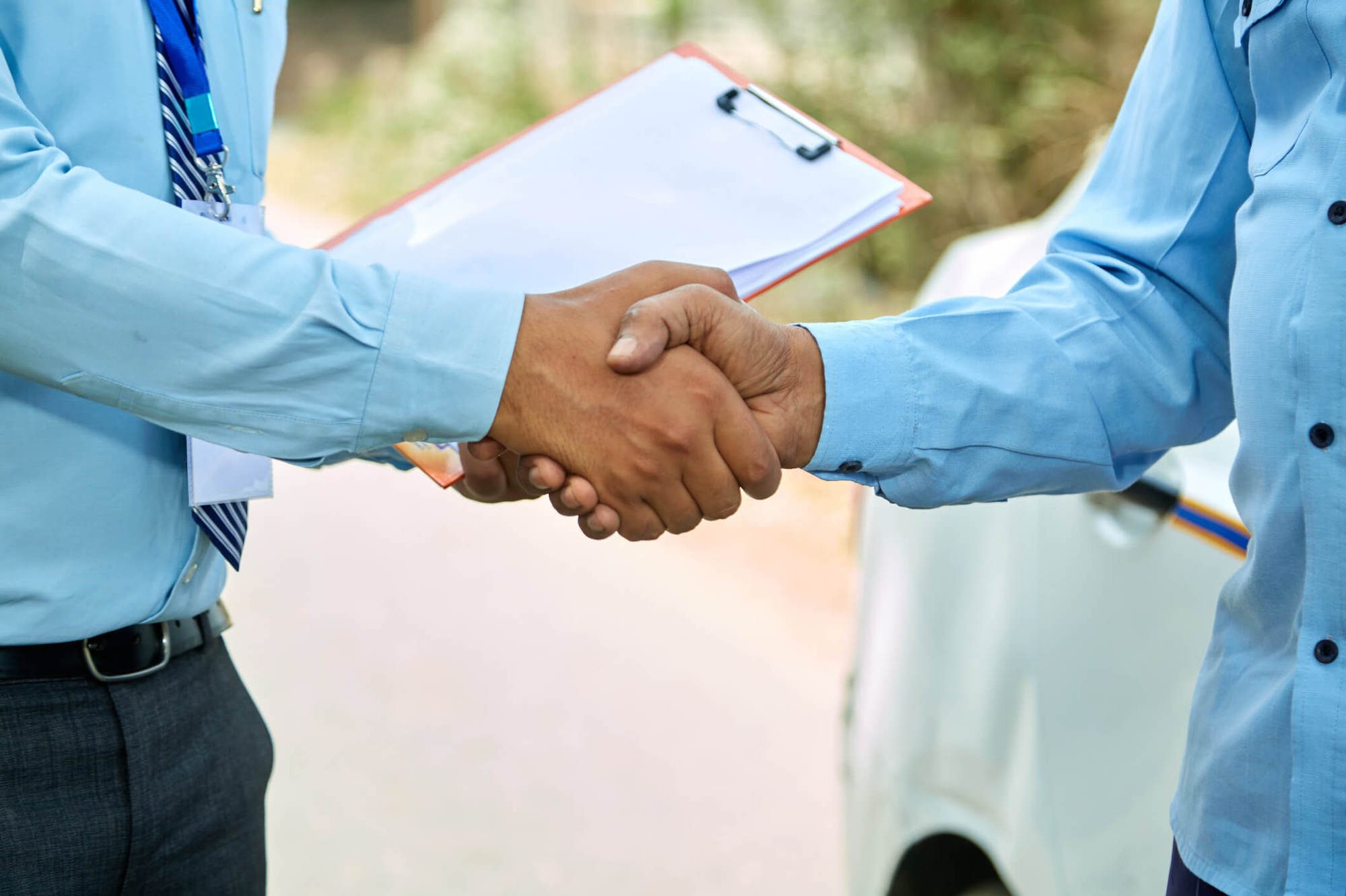 Two people in blue shirts shaking hands outdoors, with a clipboard visible in the background.