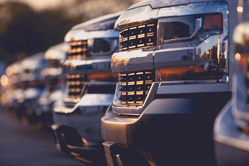 Row of silver trucks parked at sunset, with focus on the front grills and headlights reflecting warm light.